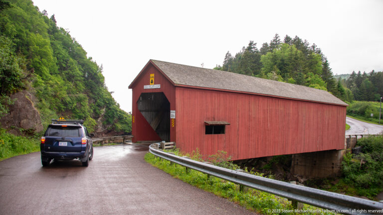Vehicle approaching the red entrance of Point Wolfe covered bridge on a wet forest road.