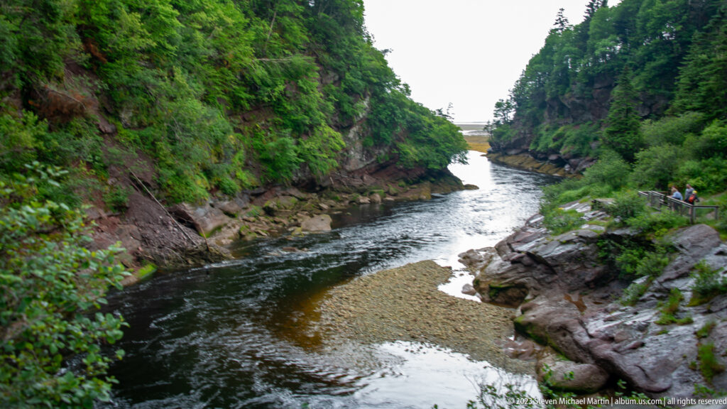 Narrow dark river flowing through a rocky forest gorge with a pale gravel bar and visitors at right.