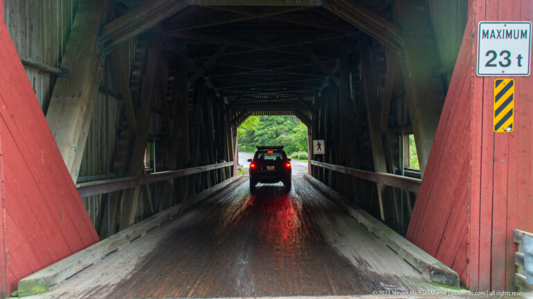 Vehicle driving through the timber interior of Point Wolfe covered bridge on wet wooden decking.