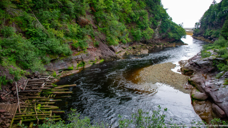 Point Wolfe River curving through a rocky gorge around a pale gravel bar with logs piled along one bank.