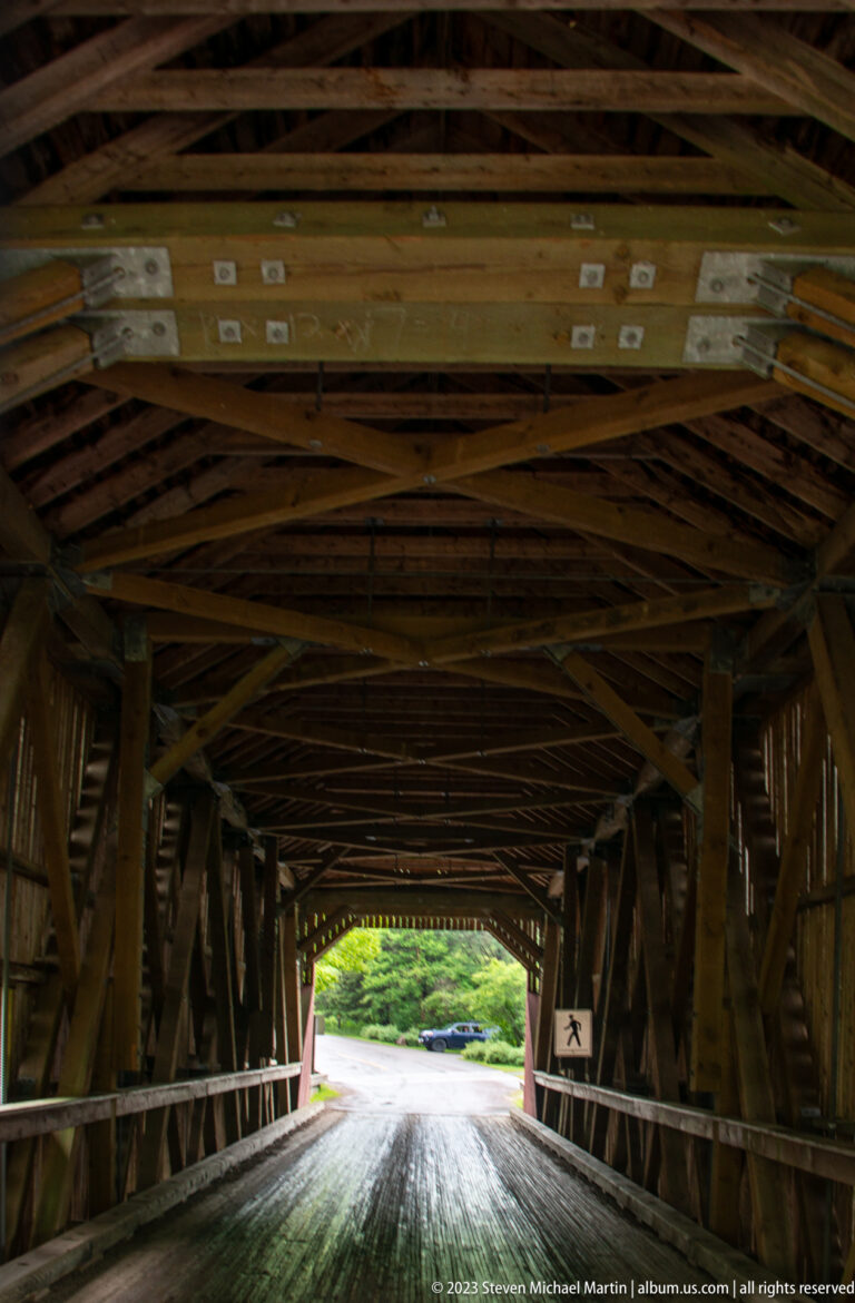 Dark timber interior of Point Wolfe covered bridge with roof beams, metal connectors, and roadway leading outside.