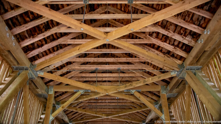 Upward view of Point Wolfe covered bridge roof with timber trusses, cross-bracing, and metal connection plates.