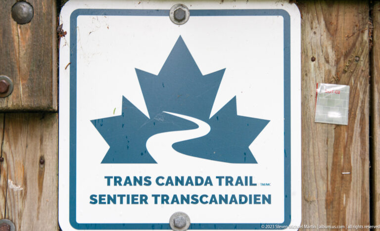 Blue-and-white bilingual Trans Canada Trail marker attached to weathered wooden boards.