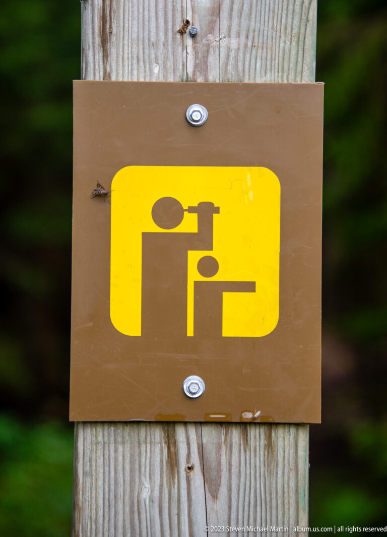 Yellow trail pictogram of a person using a viewing device, mounted on a wooden post.