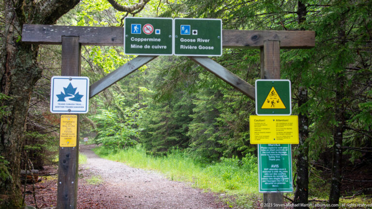 Timber trail gateway with bilingual Coppermine and Goose River signs above a gravel path into forest.