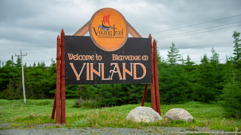 Viking Trail gateway sign reading “Welcome to Vinland” beside a spruce-lined road under cloudy skies.