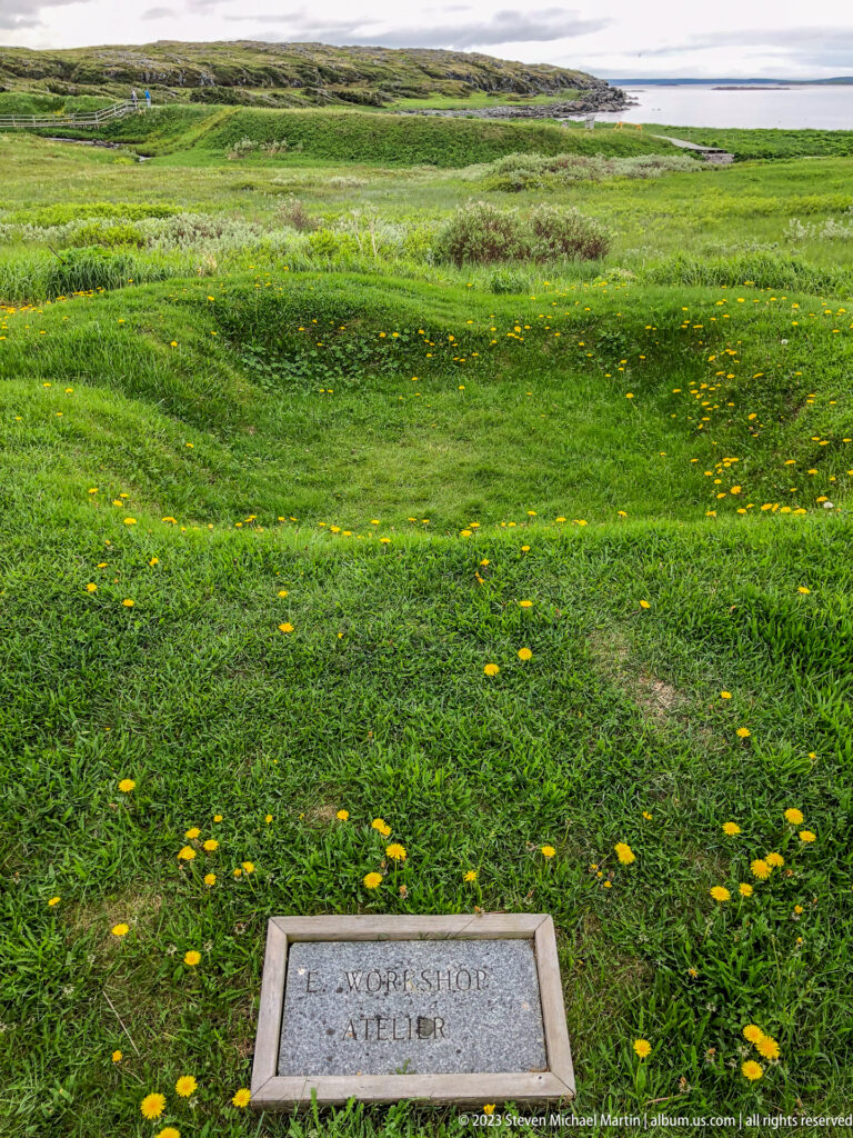 Rectangular ground marker reading Excavation and Atelier in a green coastal meadow with rocky shoreline and sea beyond.