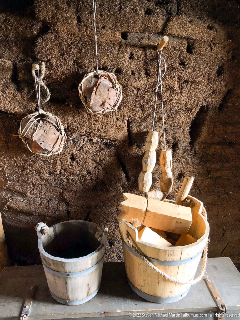 Hanging food bundles and shallow containers above wooden tubs, split wood, and a metal vessel against a dark turf wall.
