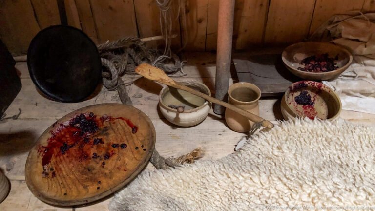 Wooden bowls with dark berries, cups, spoons, a shallow platter, and pale sheepskin arranged on a wooden surface.