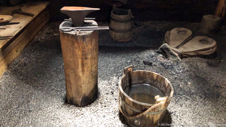 Upright wooden chopping block and water-filled wooden bucket on a gravel floor inside a dim reconstructed workshop.