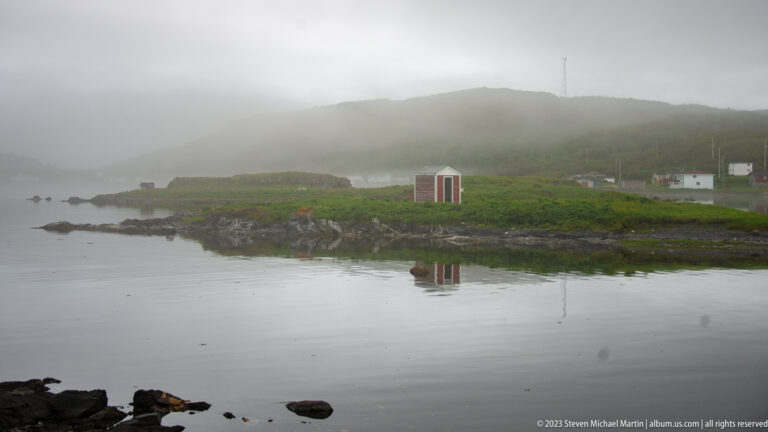 Small coastal buildings and green shoreline reflected in calm water below a broad hill partly hidden by fog.