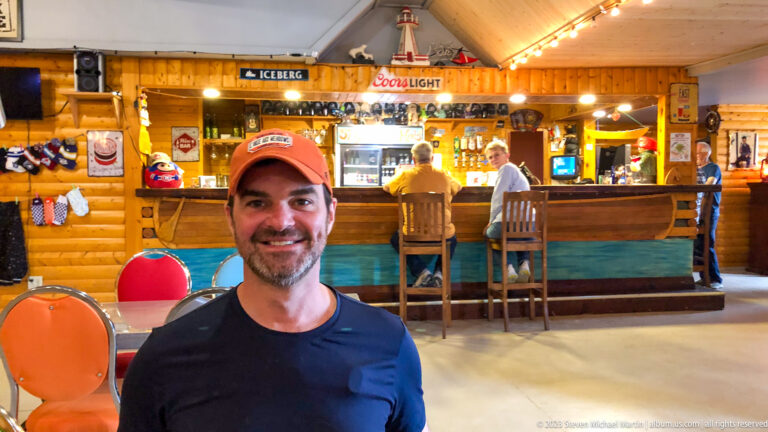 Smiling man in a red cap seated indoors with a wood-paneled bar, patrons, stools, beverage signs, and string lights behind him.