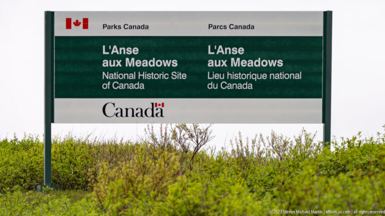 Bilingual Parks Canada sign identifying L’Anse aux Meadows National Historic Site of Canada above low shrubs.