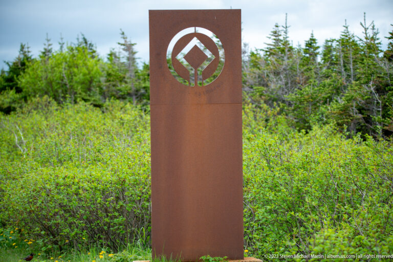 Tall rust-colored steel marker with the UNESCO World Heritage emblem standing among dense green shrubs and spruce.