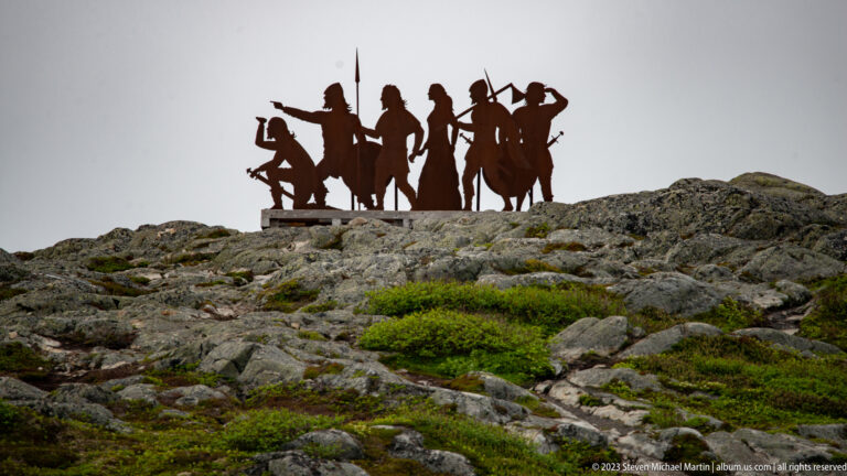 Dark metal silhouette sculpture of Viking-age figures with shields and weapons standing atop a rocky rise.