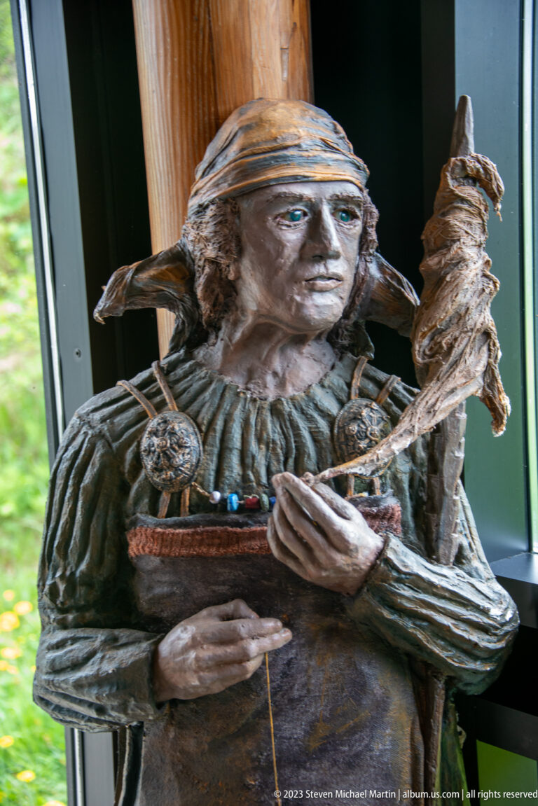 Sculpted Norse woman in layered clothing and head covering holding a fiber-wrapped textile implement.