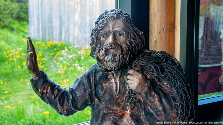 Bearded male Norse interpretive figure in a dark textured cloak, raising one hand beside a visitor-center window.