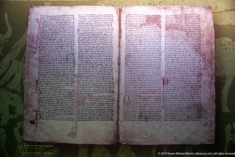 Open medieval manuscript reproduction with dense handwritten columns, colored initials, and decorative marginal marks.