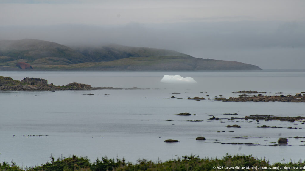 Low white iceberg on calm gray water beyond rocky shallows, with a misty headland in the distance.