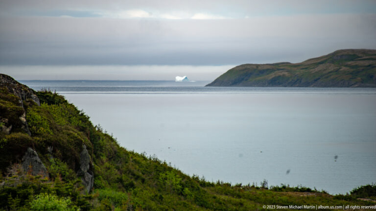 Small iceberg across a calm bay, framed by a rocky green slope at left and a long headland at right.