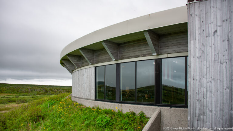 Curved wood-clad visitor centre with broad roof overhang and large windows beside a low green meadow.