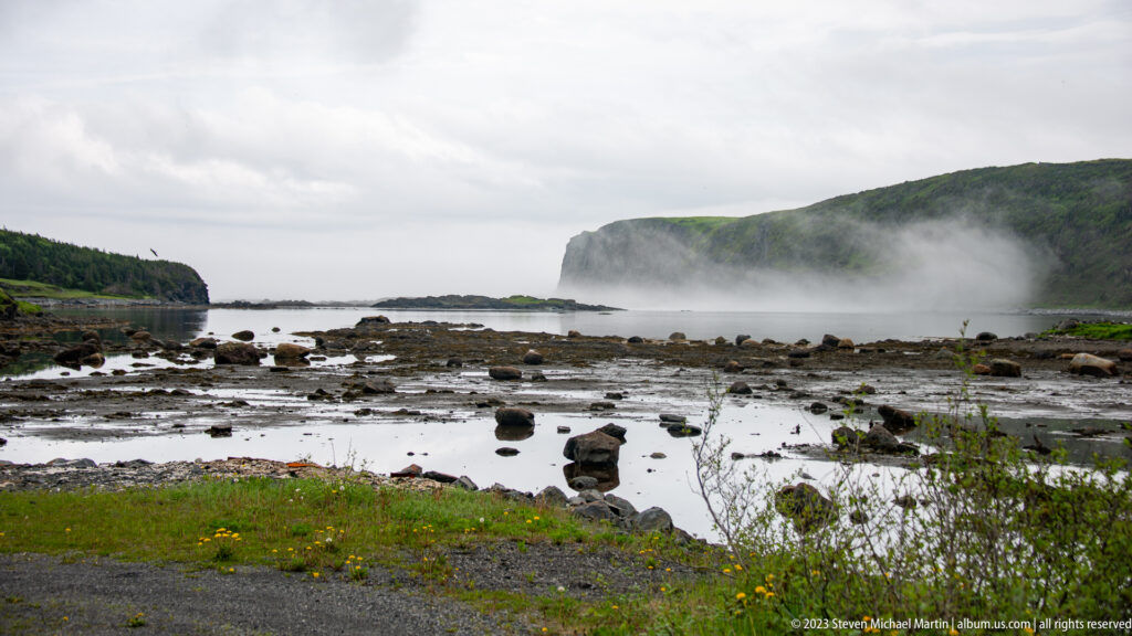 Rocky tidal flat with shallow pools and fog drifting beneath a steep green coastal headland.