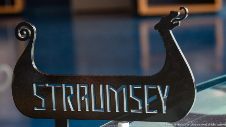 Black Viking longship-shaped metal display with curled stern, animal-headed prow, and cut-out word “STRAUMSEY.”