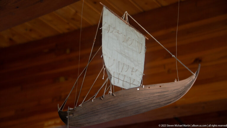 Wooden model of a Norse-style ship suspended indoors with square sail, mast, rigging, and curved clinker-built hull.