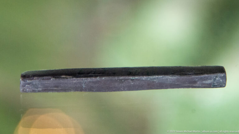 Long dark bar-shaped artifact displayed horizontally against a softly blurred green background.