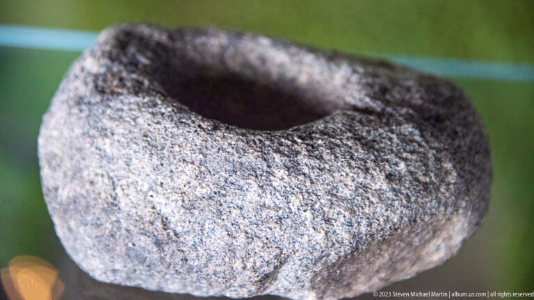 Rounded gray stone artifact with a deep hollow in its upper surface and thick coarse-textured walls.