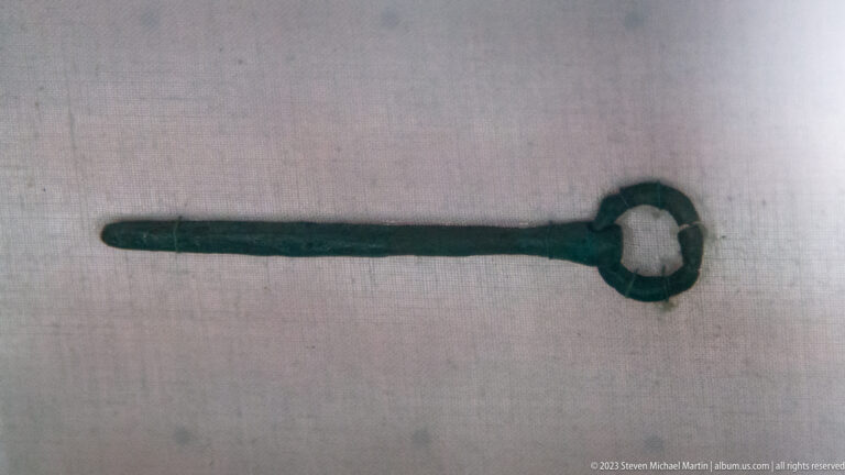 Corroded elongated metal artifact with a circular ring-shaped head resting on pale fabric.