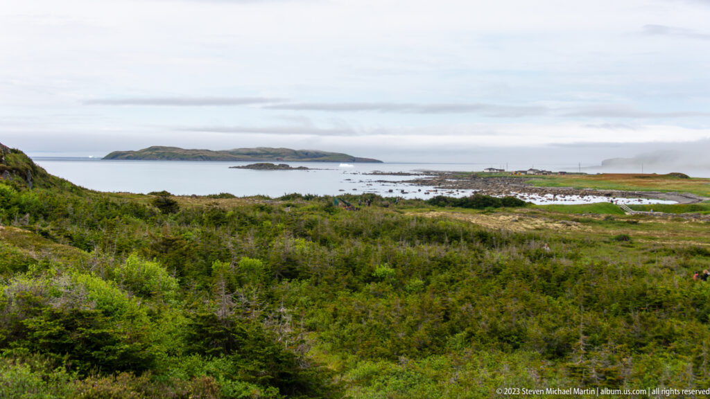 Green coastal scrub overlooking a rocky shoreline, calm sea, and low offshore island under cloudy sky.
