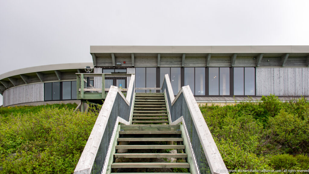 Wooden stairs rise through low green vegetation to the long glazed façade of the visitor centre.
