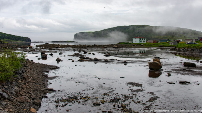 Small coastal buildings beyond a rocky tidal flat, with a fog-covered headland rising across the water.