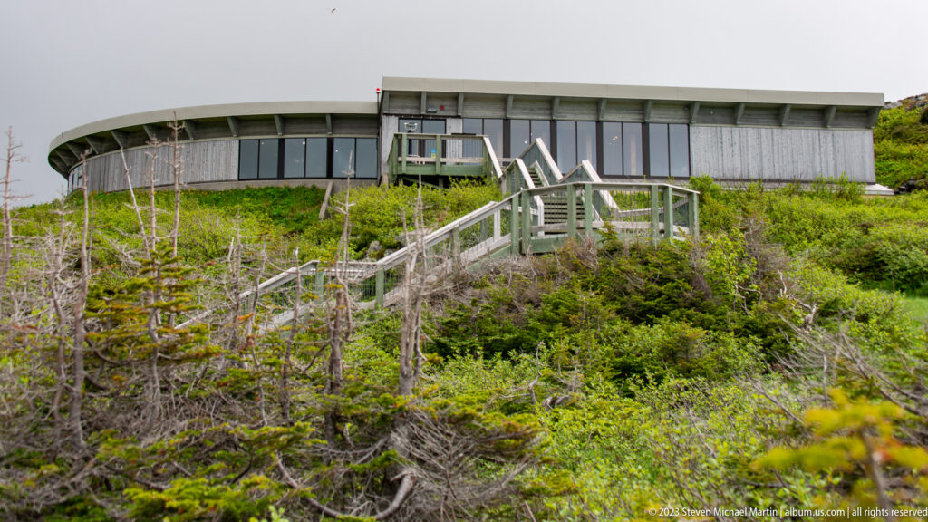Long visitor-centre building above timber stairs and railings amid spruce and dense hillside vegetation.