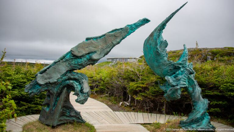 Two large curving blue-green bronze sculptural forms rise beside a wooden boardwalk amid low coastal vegetation.