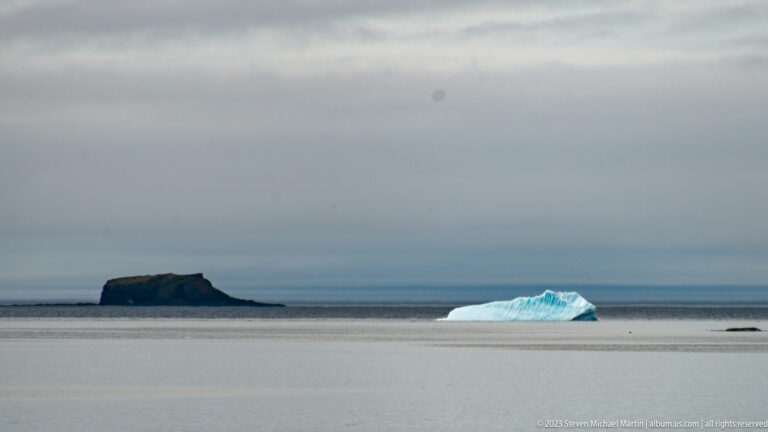 Blue iceberg floating in front of a dark flat-topped offshore islet on a calm gray sea.