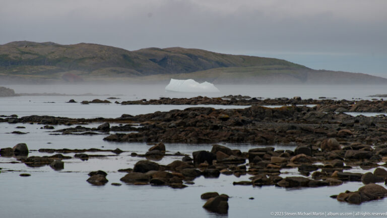 Dark boulders and tidal rocks in the foreground with a distant white iceberg and misty headland beyond.