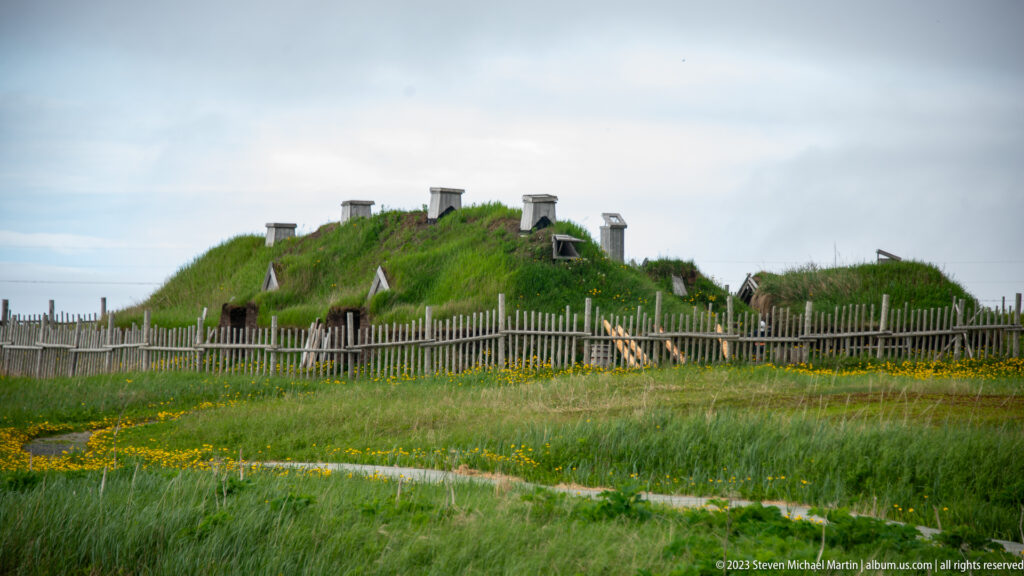Grass-covered reconstructed turf buildings with roof vents behind a wooden palisade in a green meadow.