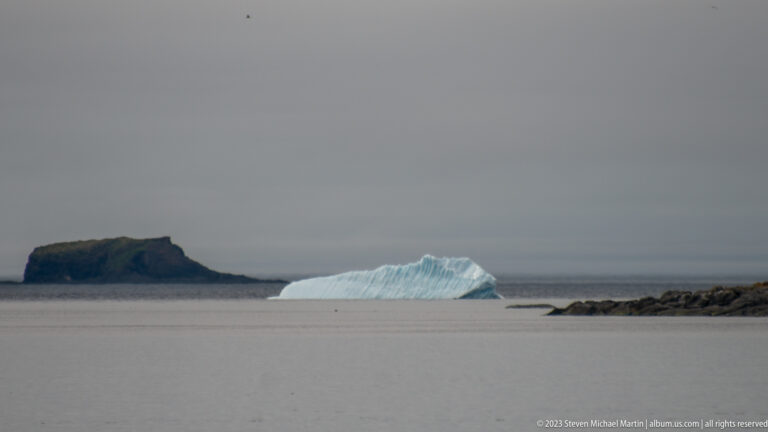 Blue-white iceberg offshore beside a dark flat-topped islet, with a rocky point entering from the right.