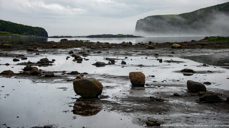 Large rounded boulders reflected in shallow tidal pools, with mist and a steep coastal headland beyond.
