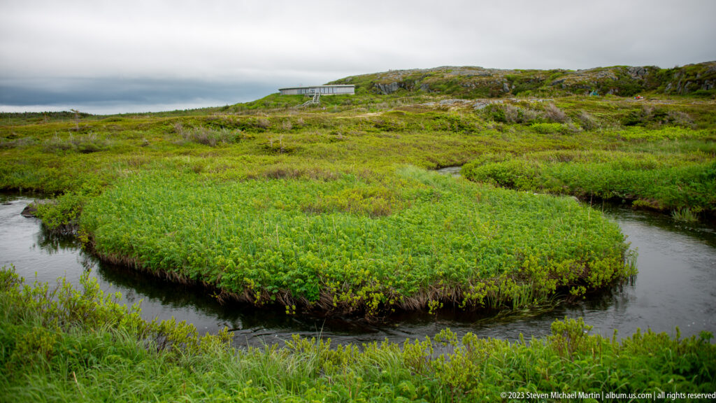 Dark stream bends around dense green wetland vegetation with low rocky hills and a distant boardwalk.