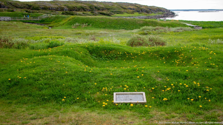 Low grassy earthworks and shallow depressions beside a small interpretive marker, with the coast visible beyond.