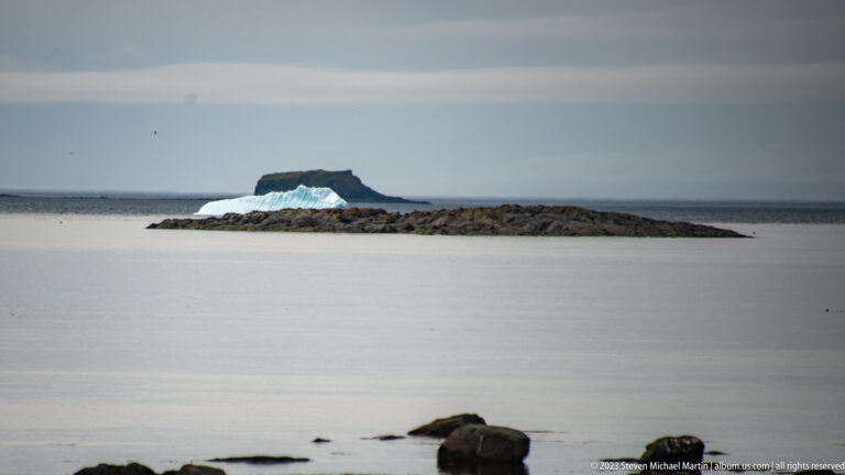 Pale blue iceberg beyond a low rocky ledge with a dark flat-topped islet on the horizon.