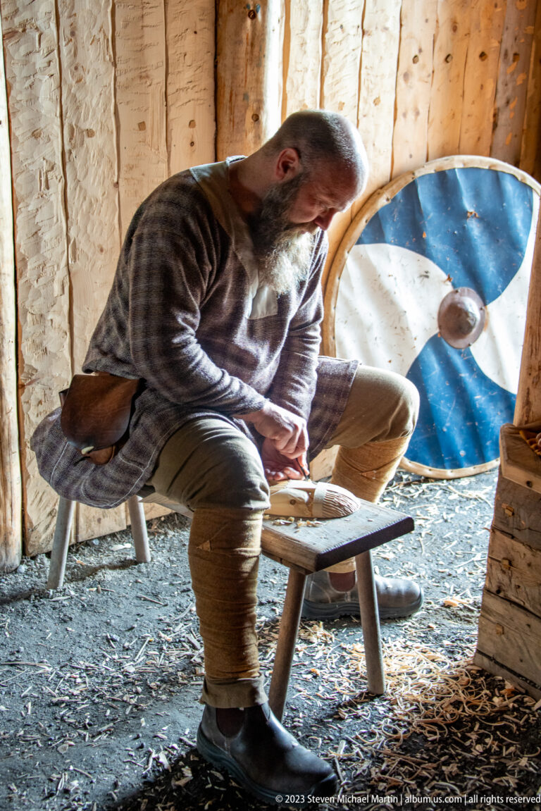 Costumed interpreter seated on a low bench inside a wooden reconstructed dwelling beside a blue-and-white round shield.