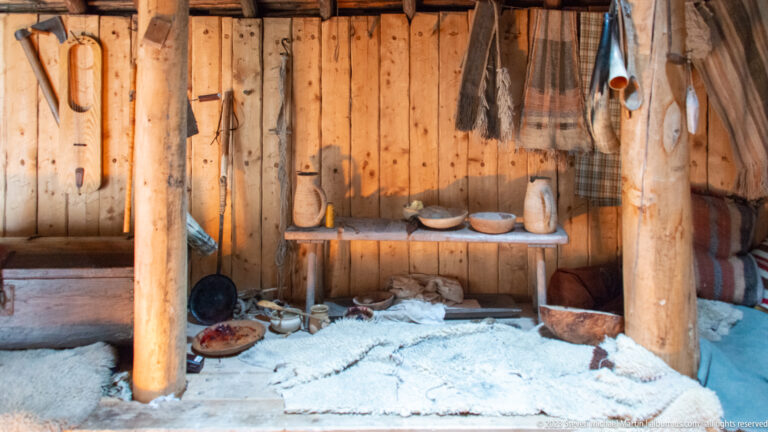 Wooden shelf with bowls and containers above pale bedding and textiles inside a rough-planked reconstructed dwelling.