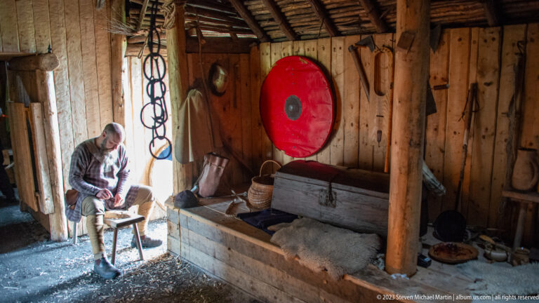 Costumed interpreter in a timber reconstructed dwelling with red round shield, wooden chest, baskets, rings, and bedding.