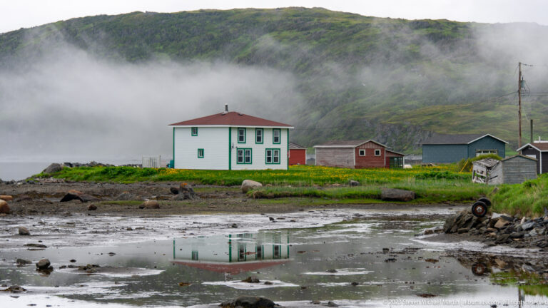 White two-story coastal house with green trim and red roof reflected in tidal water beneath a foggy hillside.