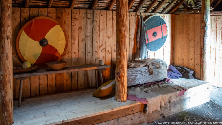 Raised bed with hides and textiles inside a timber dwelling, flanked by two brightly painted round shields.