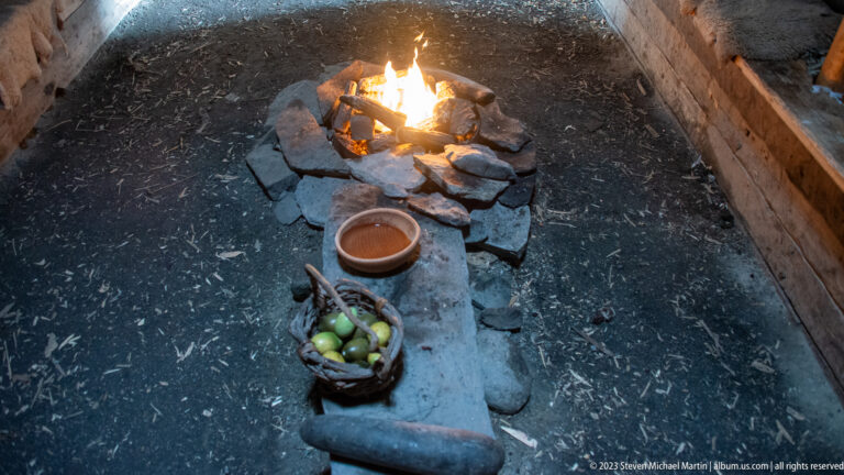 Open stone hearth with burning fire, clay bowl, and basket of green produce on an earthen floor inside a turf dwelling.